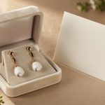 Quelles idées de cadeaux symboliques offrir pour vingt ans de mariage ? Écrin ouvert présentant des boucles d’oreilles artisanales en porcelaine blanche et or, posé près d’une carte élégante sur une table raffinée, dans une lumière douce.