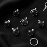 Vue de dessus de bijoux symboliques argentés ornés de pierres sombres, disposés avec élégance sur un tissu noir sous une lumière douce.